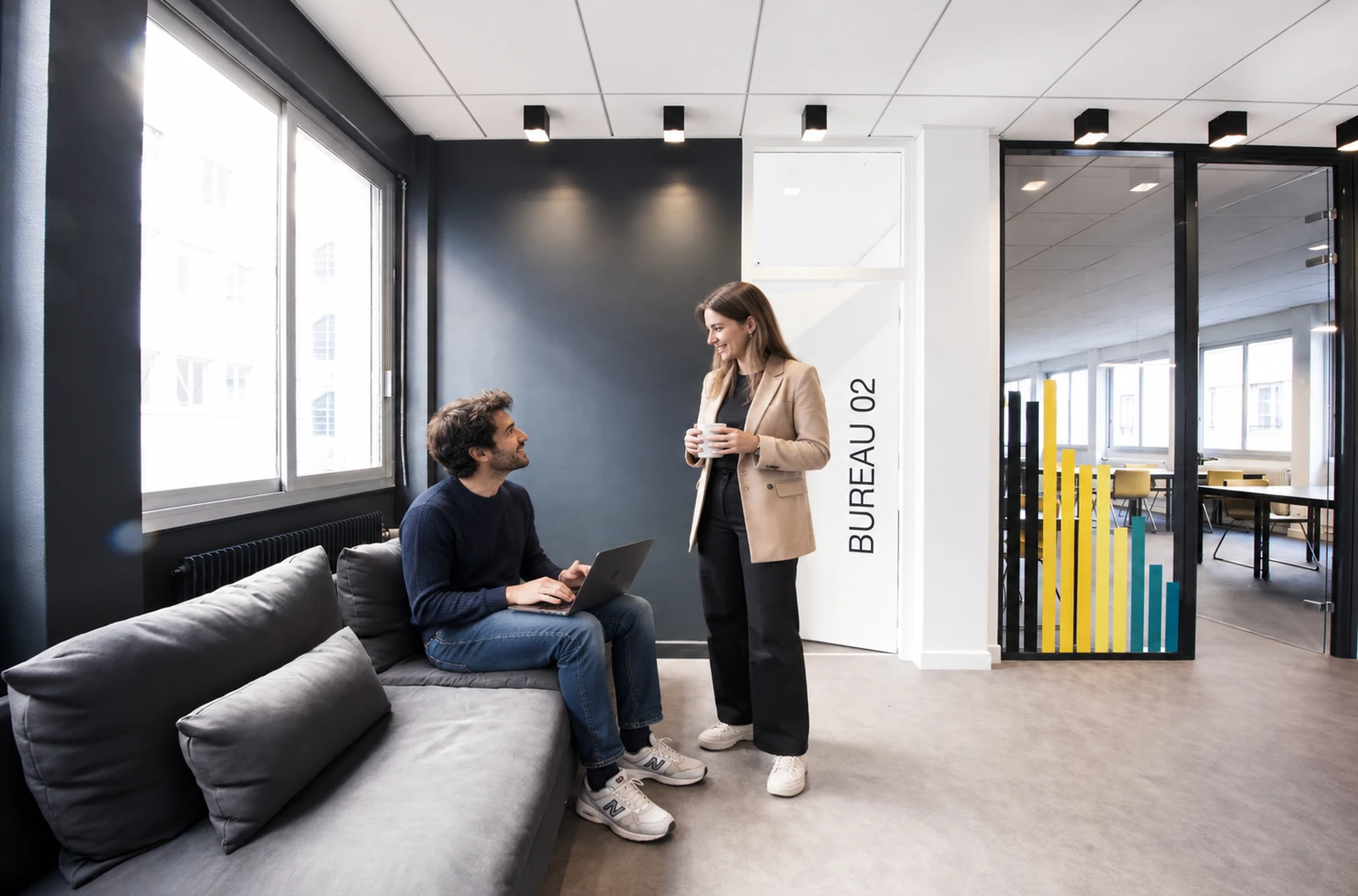 Le matin dans le salon : une coworkeuse arrive avec son café, un coworker la salue depuis le canapé.