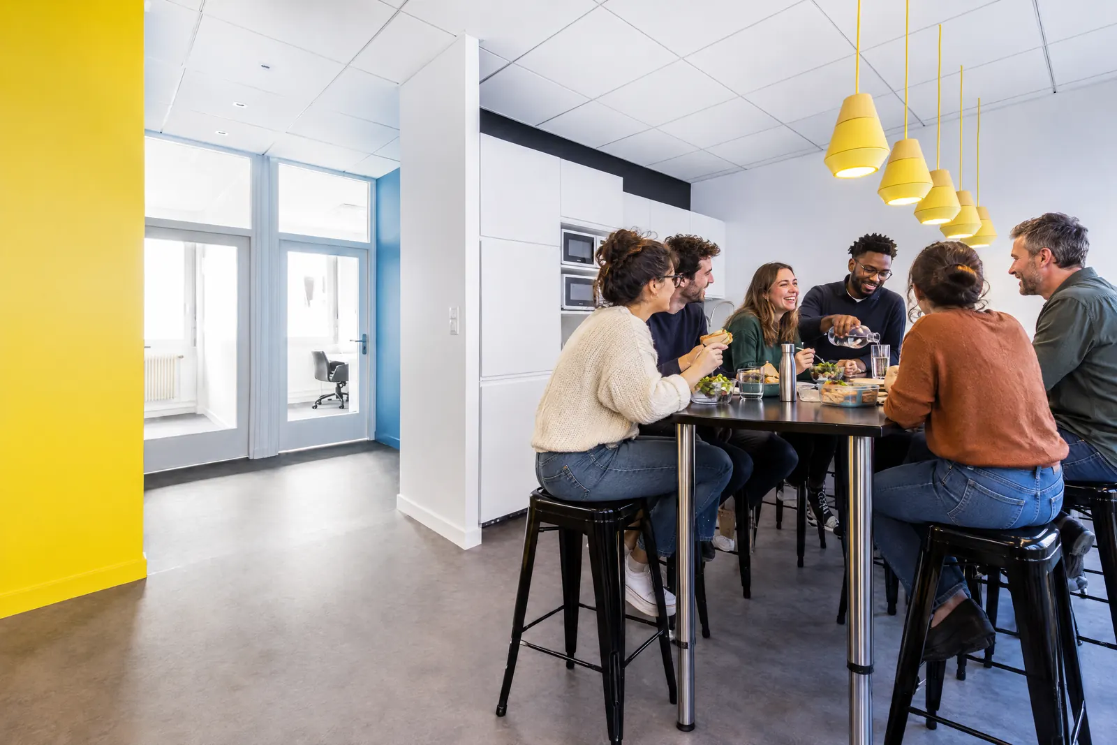 La pause déjeuner : six coworkers partagent leur repas autour de la table haute de la cuisine.