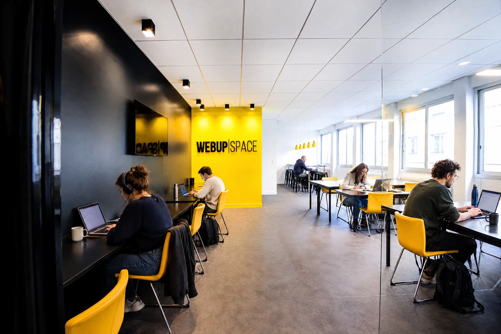 L'open space en fin de matinée : cinq coworkers concentrés sur leurs ordinateurs devant le mur jaune au logo.