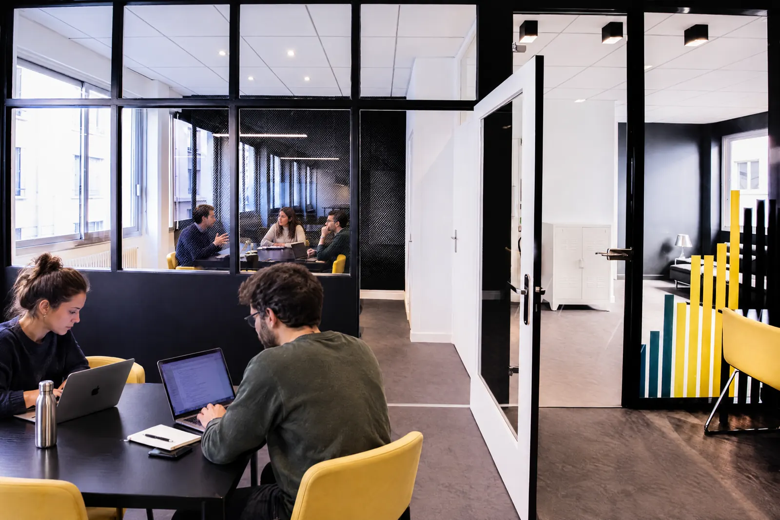 La salle de réunion vitrée : trois personnes en réunion, deux coworkers au travail dans l'open space au premier plan.